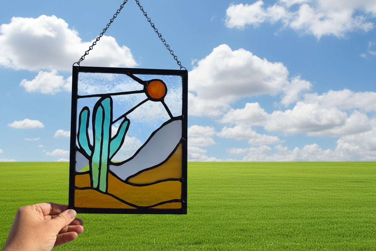 Stained glass panel featuring a bold green cactus, warm orange sun, and layered desert landscape in amber and sand toned glass, framed in dark solder lines and approximately 8.5 by 11 inches.