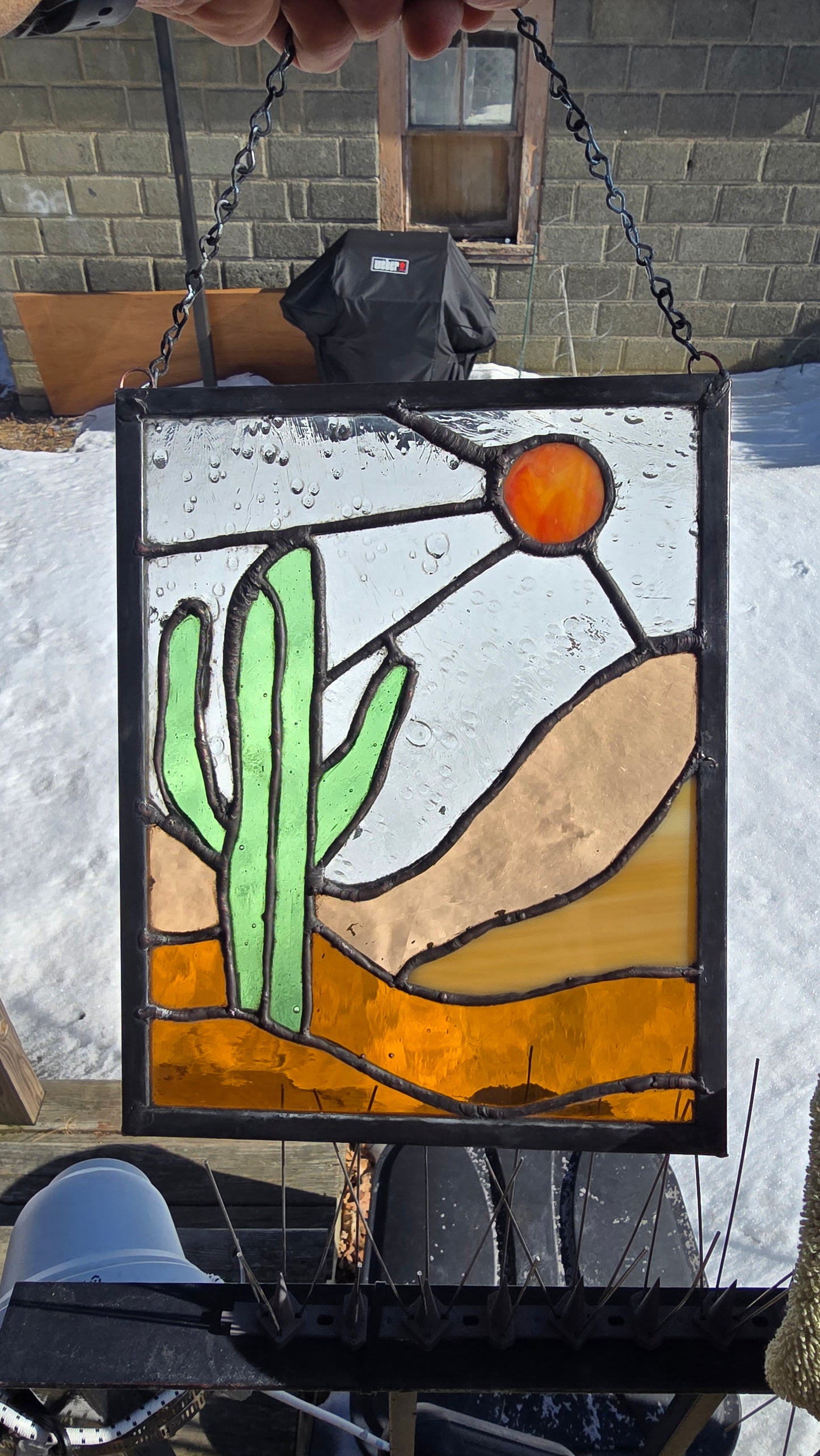Stained‑glass panel featuring a bold green cactus, warm orange sun, and layered desert landscape in amber and sand‑toned glass, framed in dark solder lines and approximately 8.5 by 11 inches.