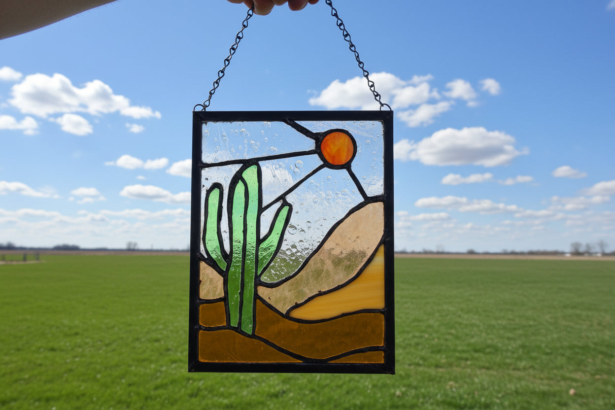 Stained glass panel featuring a bold green cactus, warm orange sun, and layered desert landscape in amber and sand toned glass, framed in dark solder lines and approximately 8.5 by 11 inches.