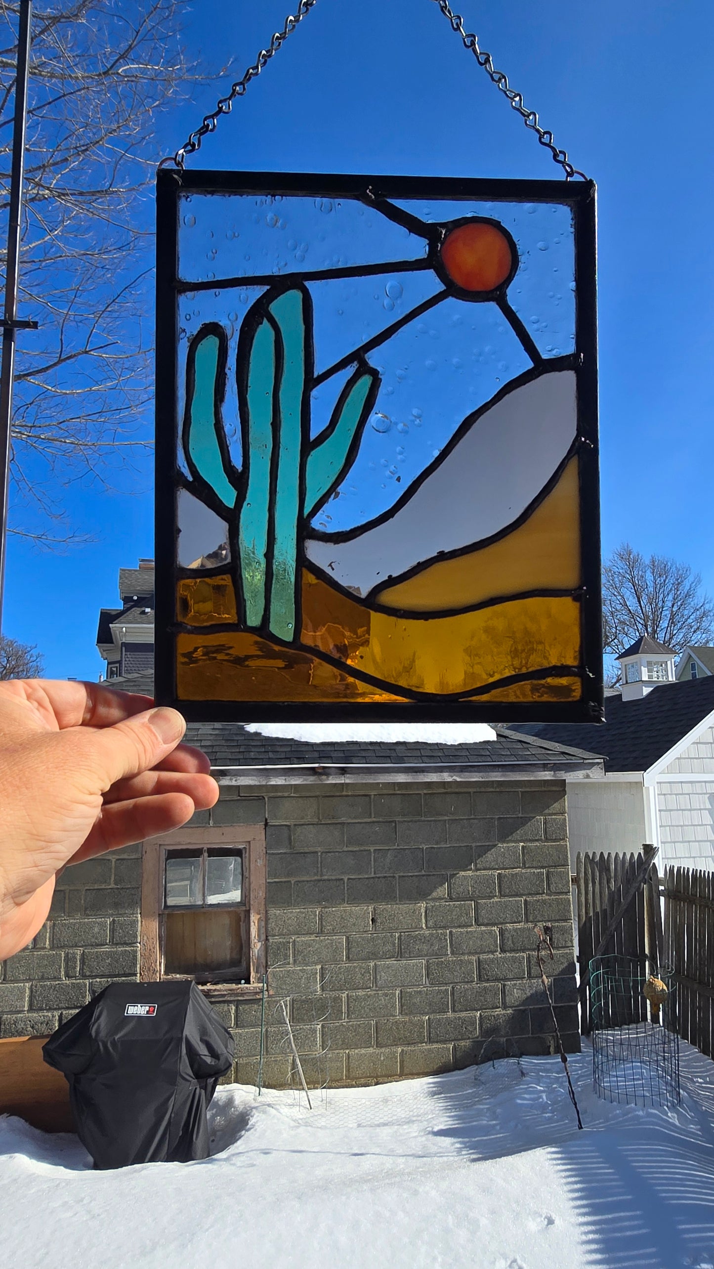 Stained‑glass panel featuring a bold green cactus, warm orange sun, and layered desert landscape in amber and sand‑toned glass, framed in dark solder lines and approximately 8.5 by 11 inches.