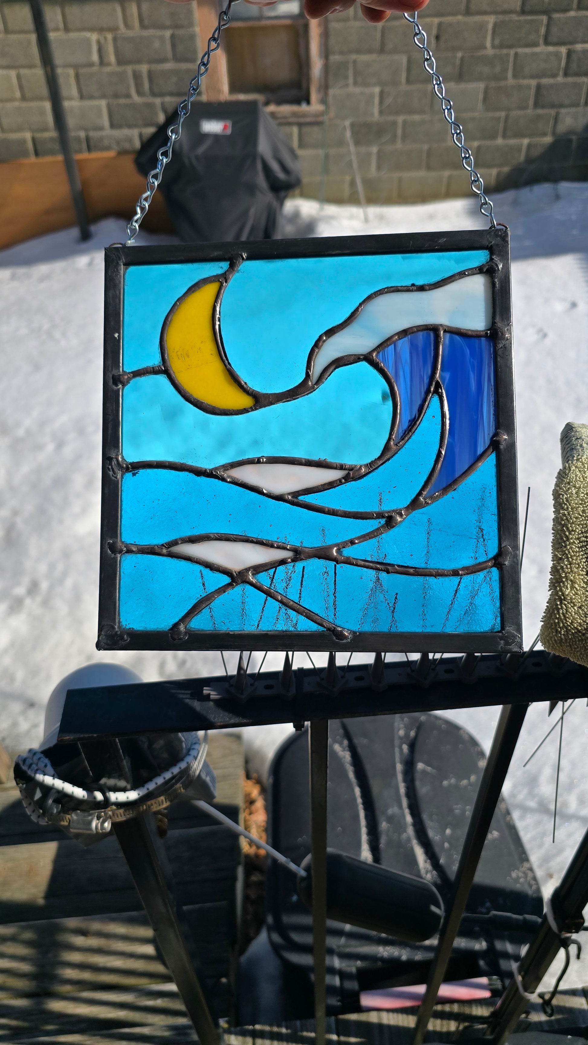 Stained‑glass panel featuring a yellow crescent moon above layered blue and white ocean waves with a sense of crashing surf, framed in dark solder and approximately 9 by 9 inches.