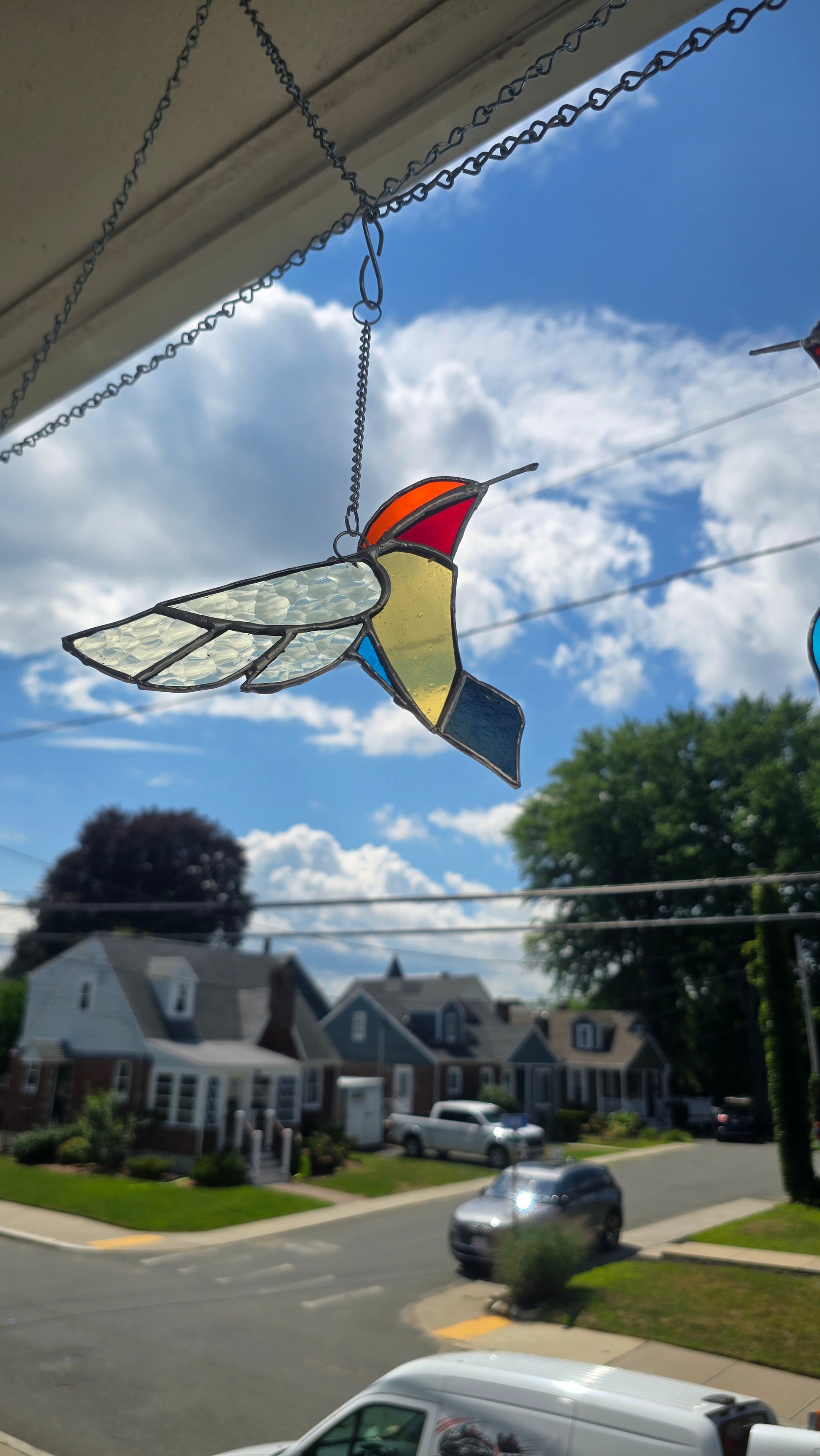 A stained glass hummingbird ornament hanging from a chain outside a house, with a suburban street and houses visible in the background under a blue sky.