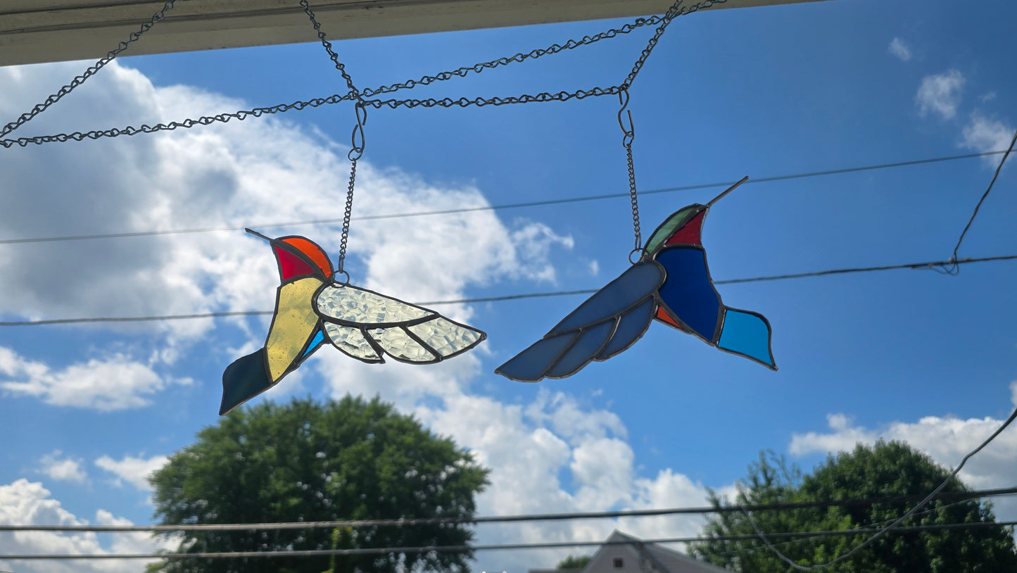 Two colorful stained glass hummingbird ornaments hanging from chains outdoors against a blue sky with clouds and trees below.
