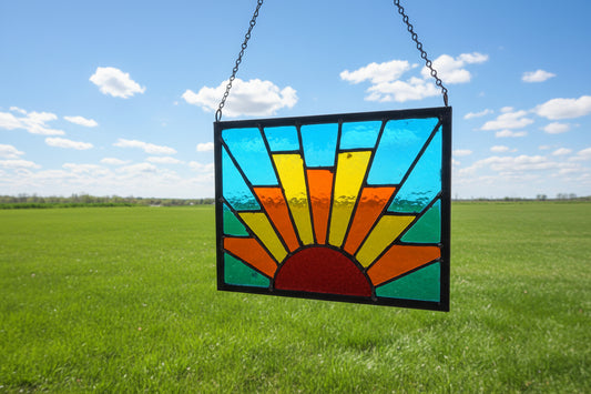 Handcrafted stained glass sun panel with bold red, orange, and yellow rays against a deep blue background, framed in dark solder lines, approximately 8.5 by 11 inches.